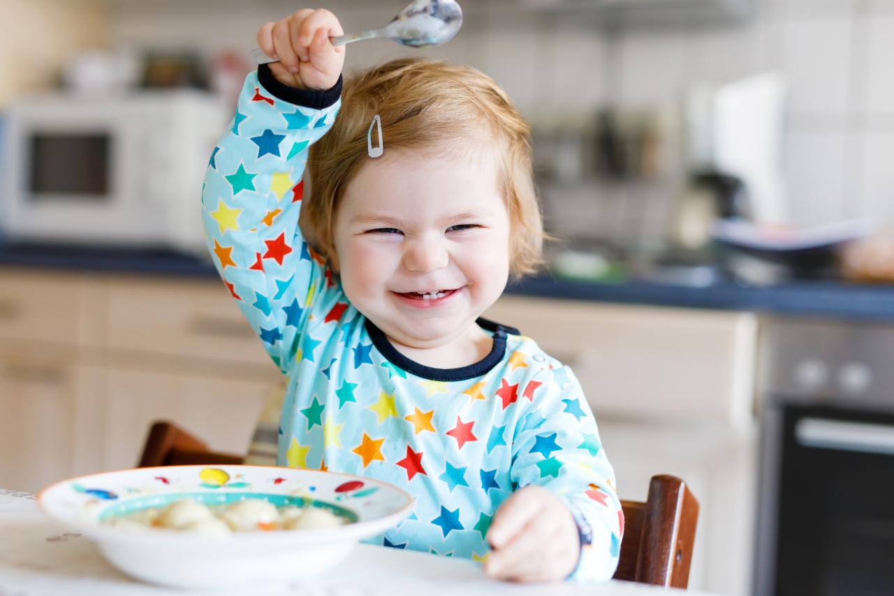 a    smiling baby  holding up a  spoon  with  bowl of food in  front of  them.