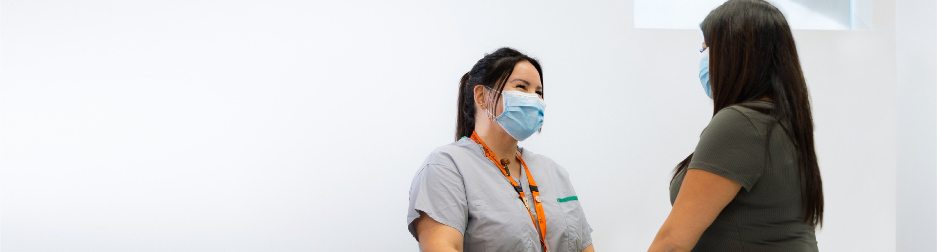 A registered nurse in a mask talks with a patient in a white room