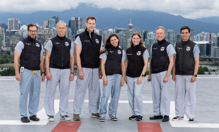 7 physicians wearing blue medical scrubs and black vests standing on a hospital rooftop with downtown Vancouver skyline and mountains in the background