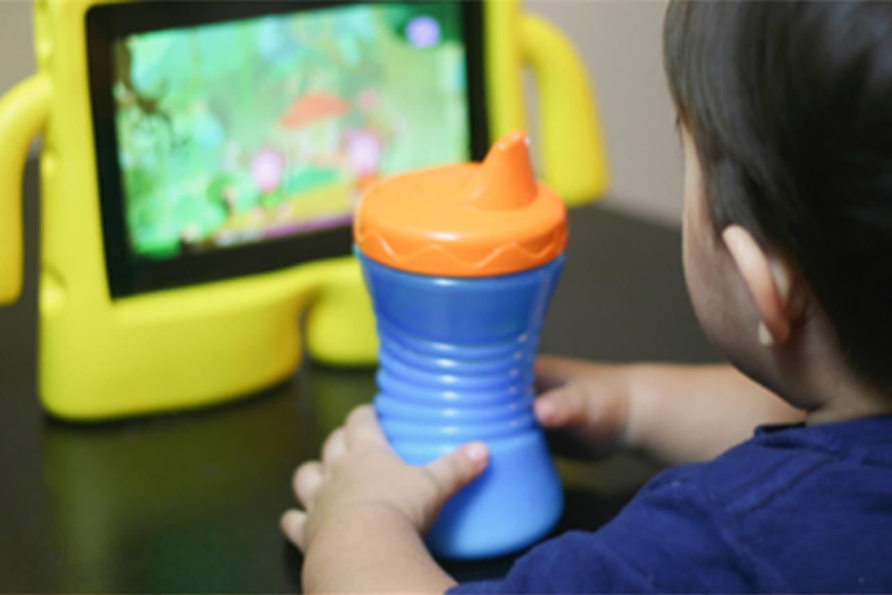 A toddler in blue shirt with a blue and orange drinking cup in front of a tablet with a yellow case