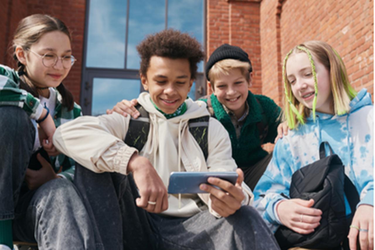 a group of four teens sitting outside a brick building looking at a smartphone screen