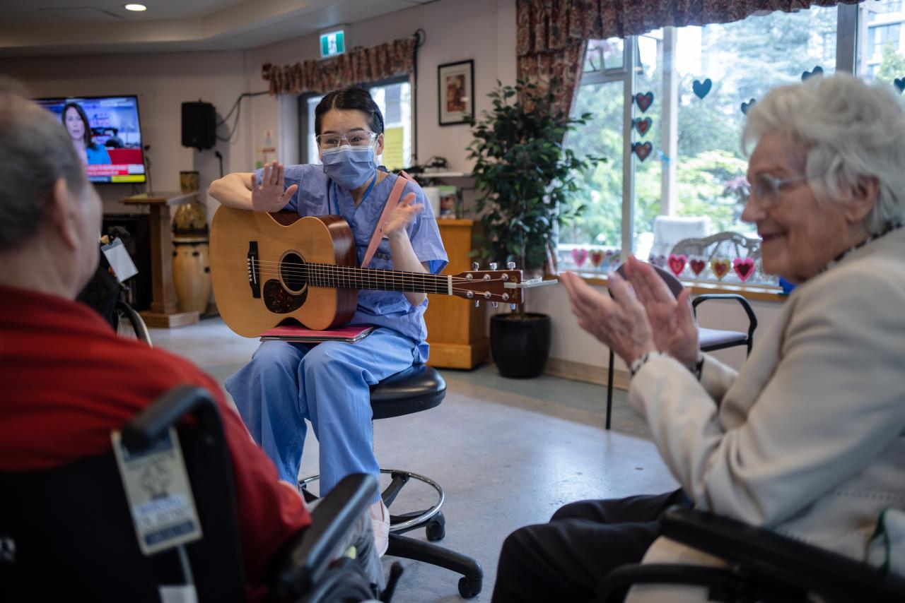 A healthcare worker wearing scrubs performs with a guitar for two elderly folks in a care home communal area