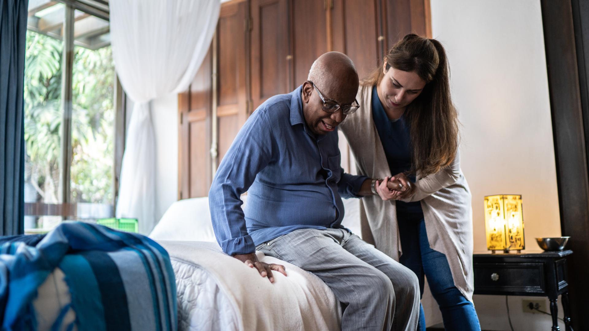 Nurse in assisted living home helps an older adult stand up from a bed