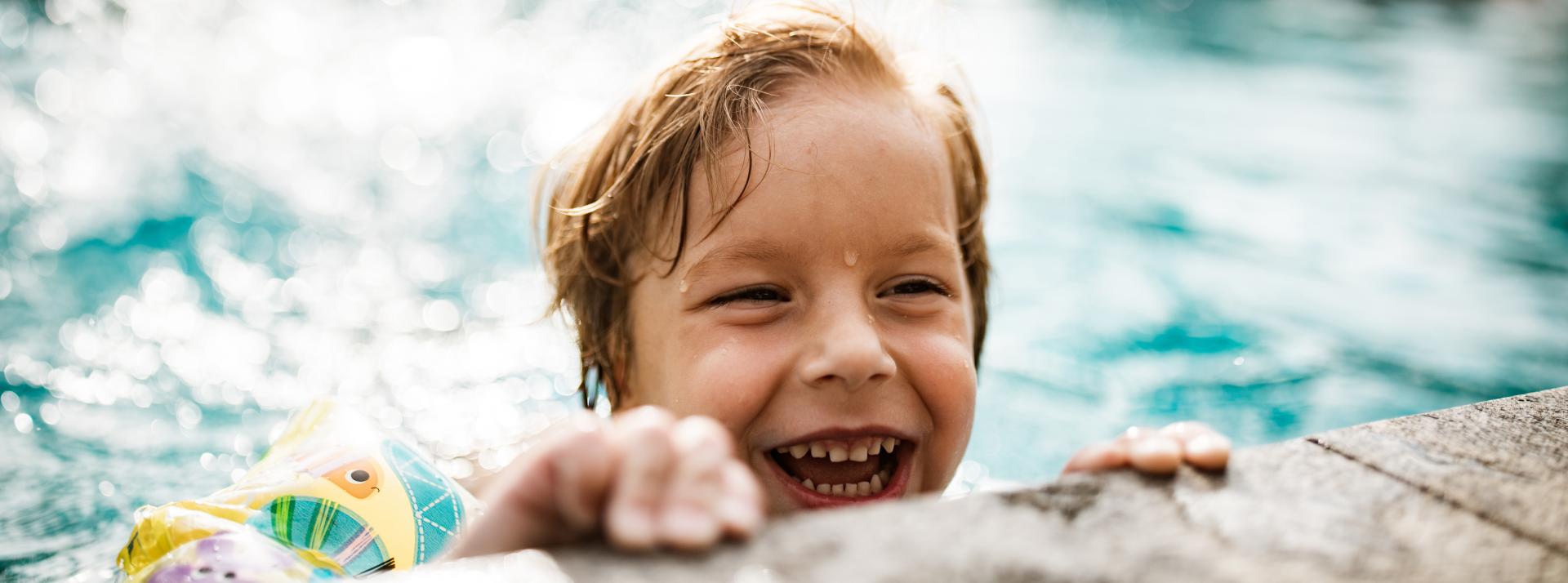 A happy child wearing water wings holding the edge of an outdoor pool on a sunny day.