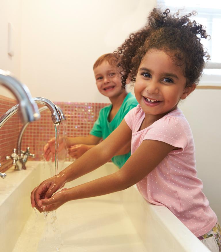 A young girl in a pink shirt smiling and washing her hands at school