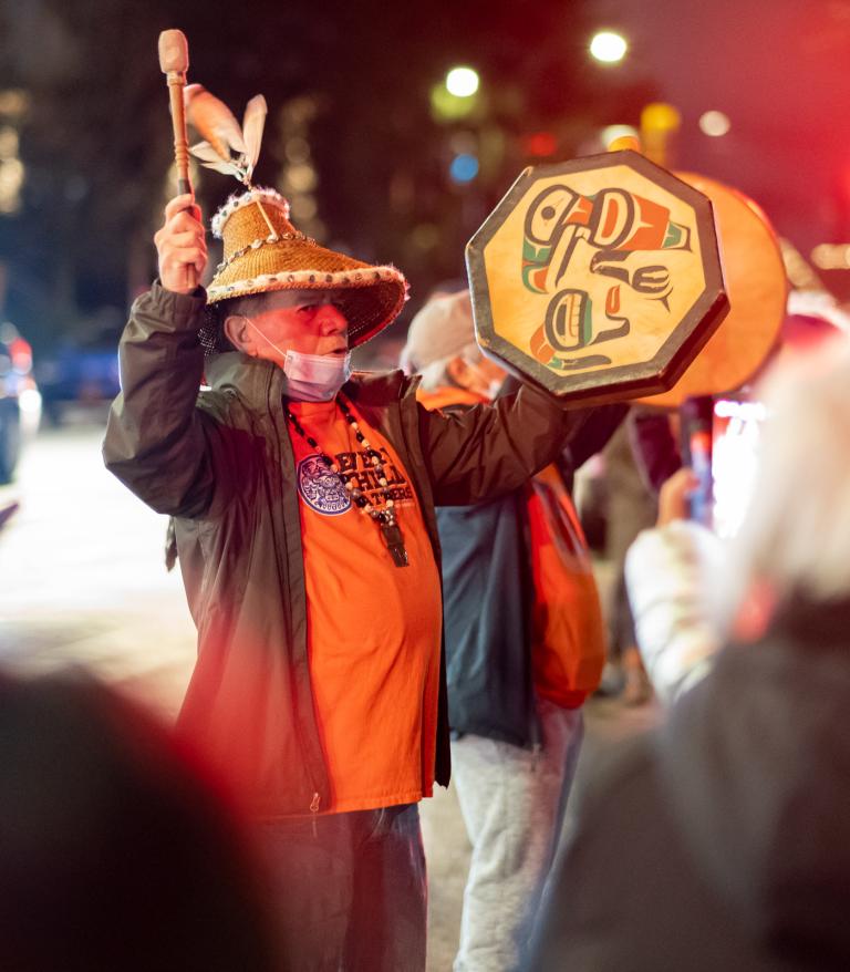 Man wearing a cedar hat and a orange shirt playing a traditional indigenous drum