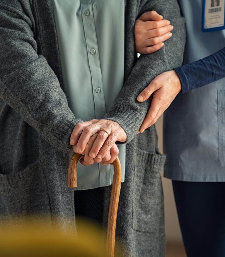 A closeup of a nurse helping an older person with a cane walk down a hallfway