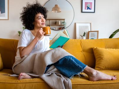 Pensive relaxed African american woman reading a book at home, drinking coffee sitting on the couch